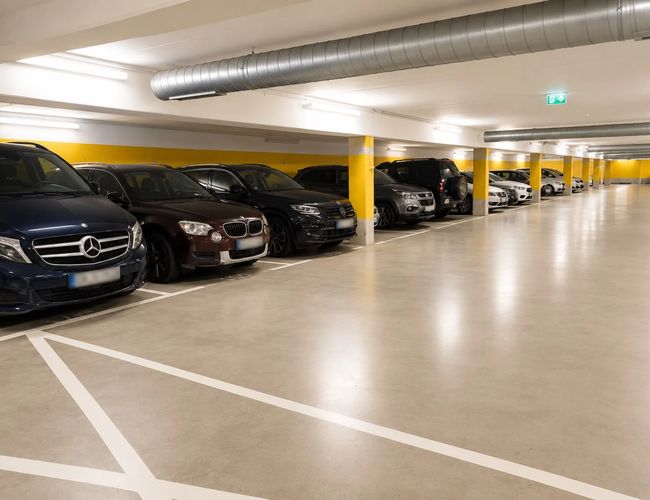 Podziemny parking z zaparkowanymi samochodami wzdłuż ściany, dobrze oświetlony przestronny korytarz.