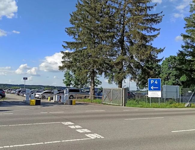 Widok na wjazd na teren parkingu od strony ulicy. W pobliżu znajdują się znaki informacyjne oraz drzewa.