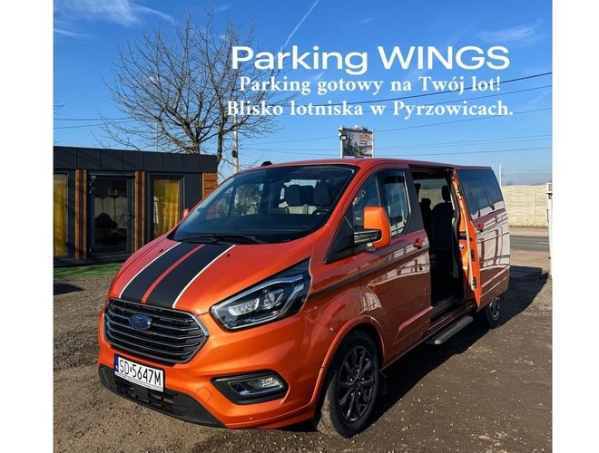 Parking Wings – pomarańczowy van stoi na placu. Teren jest ogrodzony, niebo bezchmurne. Obok widać budynek.