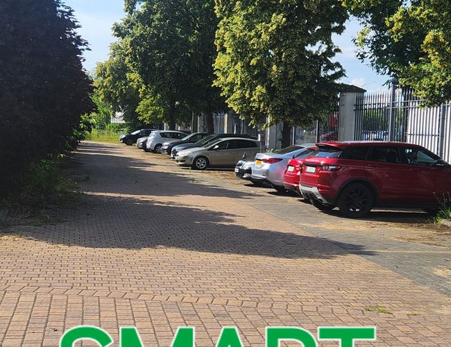 Parking Barato y Seguro SMART - los coches están alineados a lo largo de la valla. Junto a ellos crecen filas de árboles, y el terreno está pavimentado con adoquines.