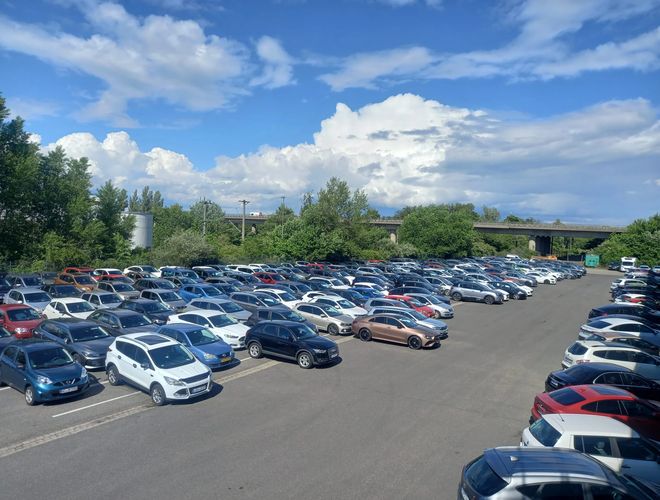 Duży parking pełen samochodów osobowych pod błękitnym niebem z nielicznymi chmurami, w tle drzewa i budynki.