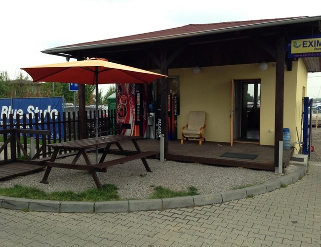 Zbliżenie na mały budynek biura parkingu, przy którym znajdują się stoliki z ławkami i parasol przeciwsłoneczny.