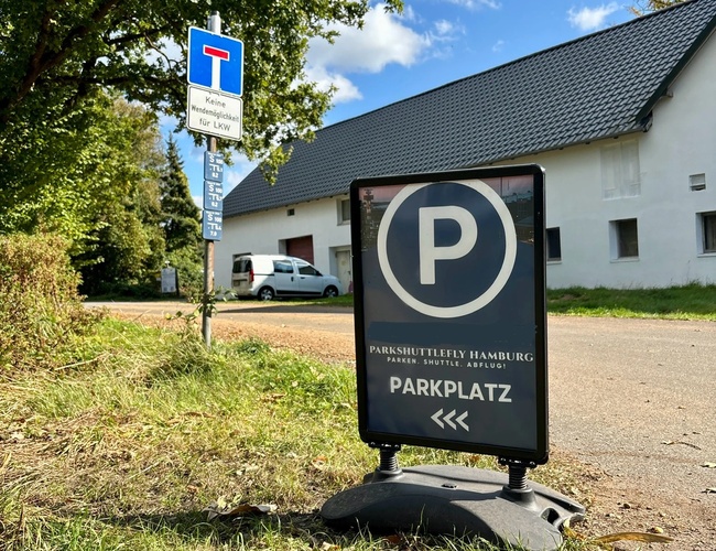 W centrum znak parking, w tle zaparkowany samochód budynek i drzewa.