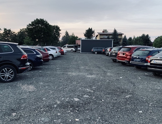 Balice Premium Park - Automobily stoja na neupravenom povrchu. Parkovisko je obklopené stromami. V pozadí je vidieť budovu.