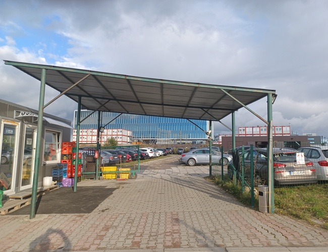 Maxi Parking – Entrada de estacionamiento cubierta y visible. Al lado hay una cerca de malla. En el fondo hay coches estacionados.