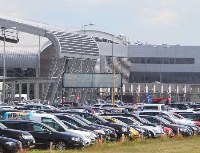 VALET Parking – wiele samochodów stoi na otwartym placu. W tle widać nowoczesny budynek lotniska. Parking jest na zewnątrz.