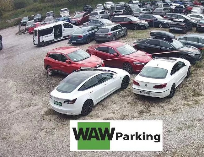 WAW Parking - Na kamenistom placu parkuje veľa áut. Priestor je plný rôznofarebných vozidiel. Na pozadí je vidieť budovu.