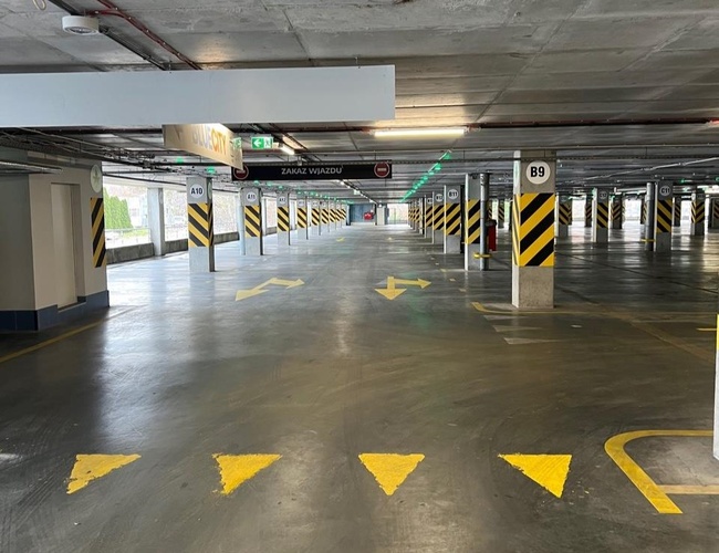 Parkovisko P2 - Okęcie Sever Viacpodlažné – priestrané parkovisko s označeniami podlaží. Betónové stĺpy majú žlto-čierne pásy. Svetlé stropné osvetlenie.