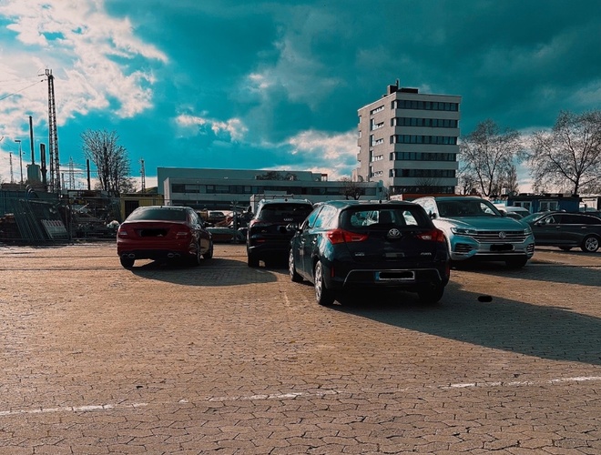 Parkplatzsicherheitparking24 VALET - Es sind einige geparkte Autos zu sehen. Im Hintergrund steht ein hoher Gebäudekomplex. Auf dem Parkplatz ist eine gepflasterte Oberfläche.
