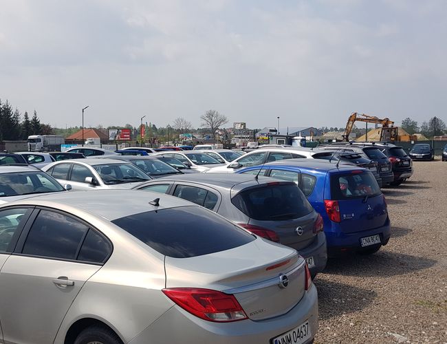 BaliceLot24 - Na parkingu stoją zaparkowane samochody. Teren jest żwirowy i ogrodzony. Po bokach widać latarnie.
