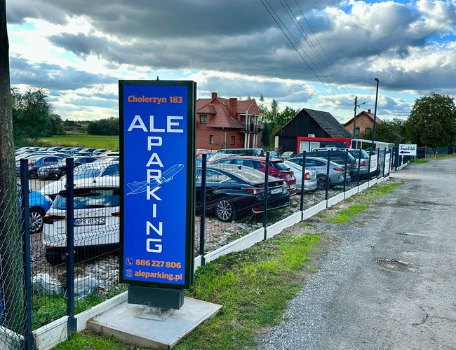 AleParking – Parking jest ogrodzony niskim płotem. Teren jest otwarty i ma żwirową nawierzchnię. Widać kilka zaparkowanych samochodów.