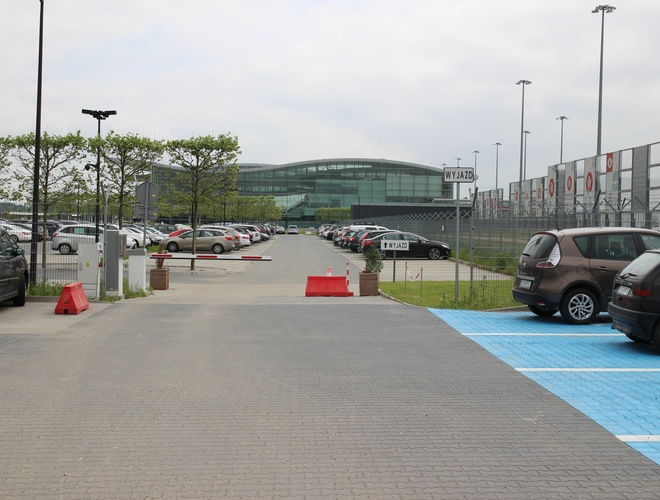 Aparcamiento Oficial Aeropuerto Wrocław Strachowice - aparcamiento con muchos coches aparcados. Aeropuerto techado al fondo. Iluminación y barrera en la entrada.