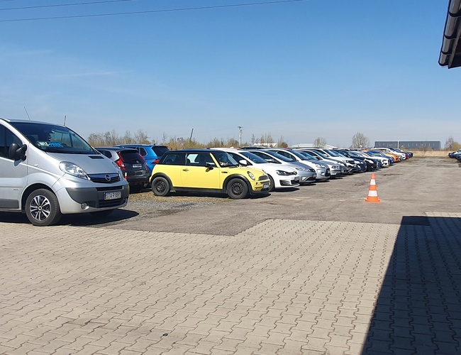 WIZ-PARK - Estacionamiento con una fila de coches a la derecha. En primer plano hay una furgoneta gris. A la izquierda hay una señal de alto.